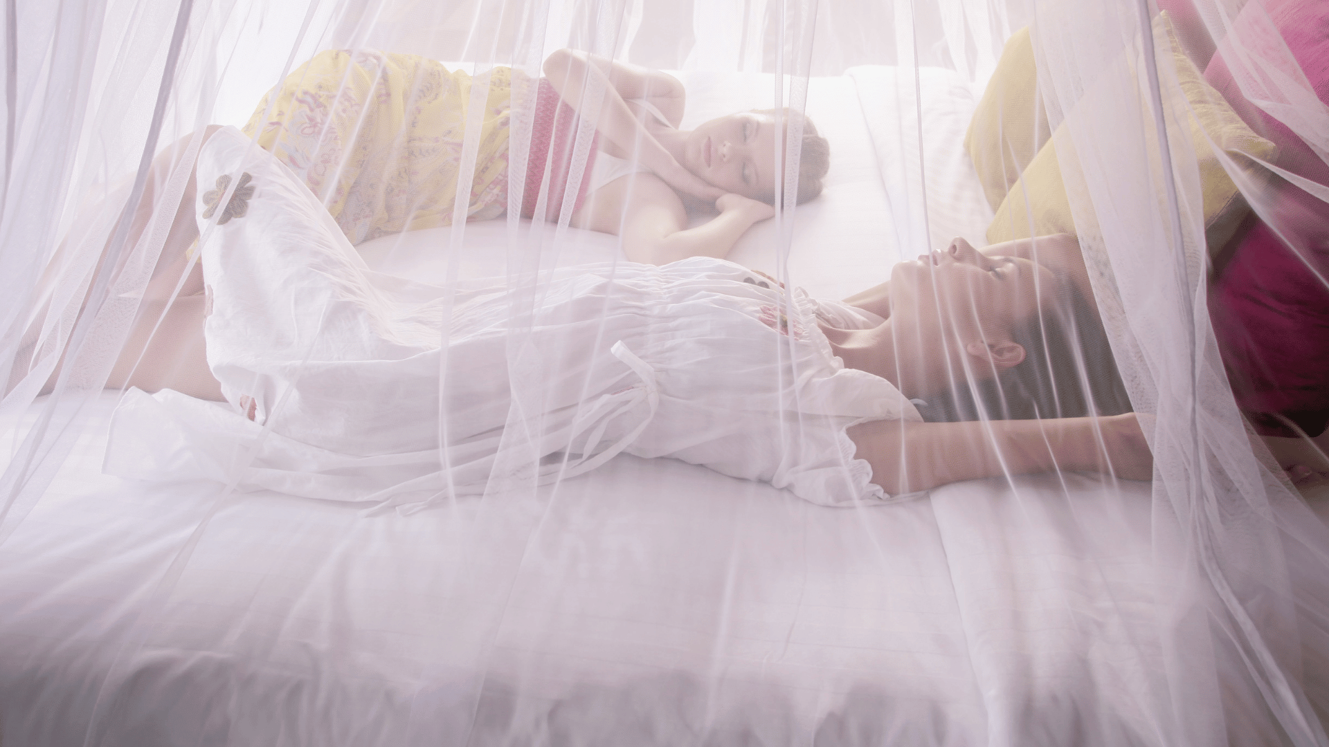 Two women resting on a white bed under a sheer mosquito net canopy, with soft natural lighting creating a calm and dreamy atmosphere.