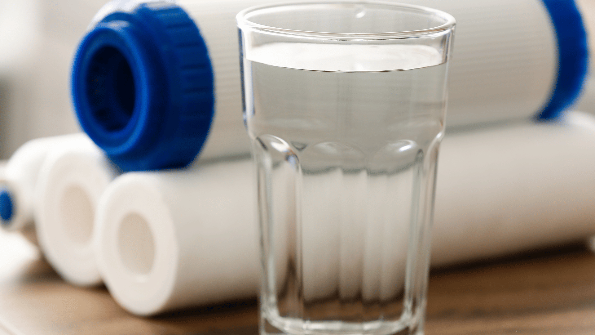 A clear glass of water sits on a wooden surface in front of several cylindrical water filter cartridges with white bodies and blue caps, suggesting a focus on clean, filtered drinking water.