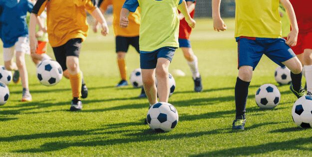 Group of children playing football on a grassy field, each wearing colorful jerseys and kicking soccer balls.