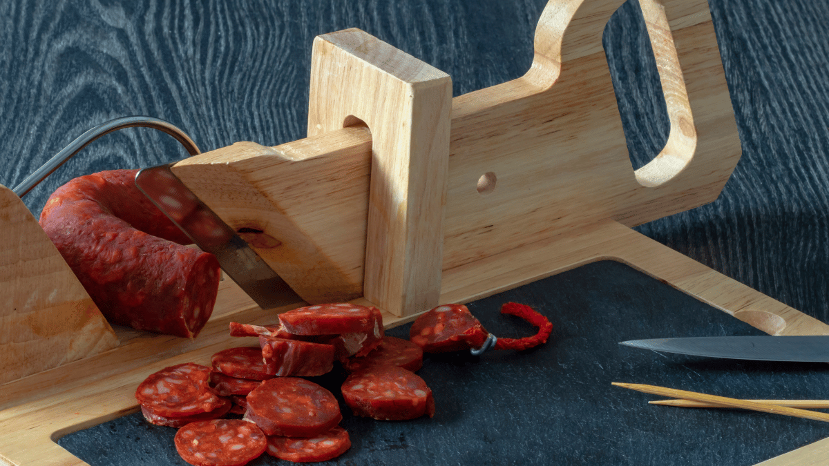 A traditional wooden sausage slicer with a partially sliced cured sausage and neatly cut pieces on a black slate board.