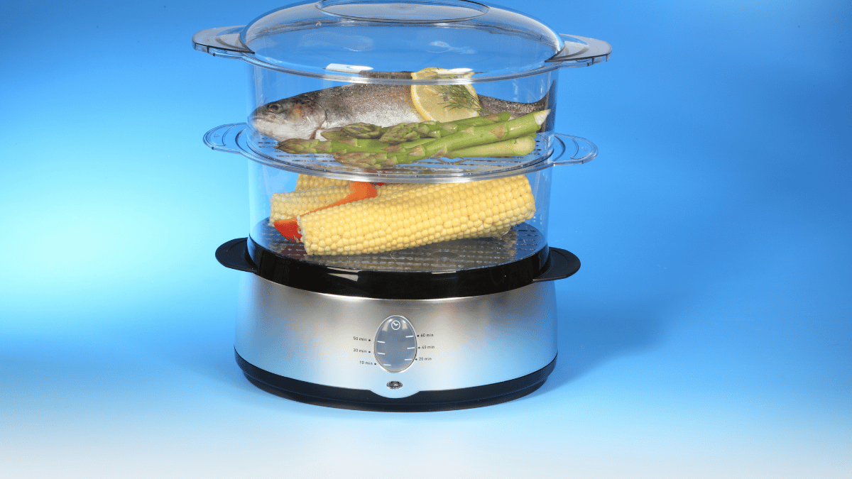 A two-tier electric food steamer cooking a whole fish with asparagus and lemon on the top tray, and corn on the cob with bell peppers on the bottom tray, against a blue background.