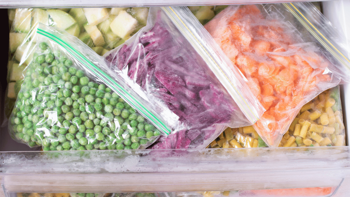 Close-up of a freezer drawer filled with sealed plastic bags of assorted frozen vegetables, including peas, carrots, beets, zucchini, and green beans