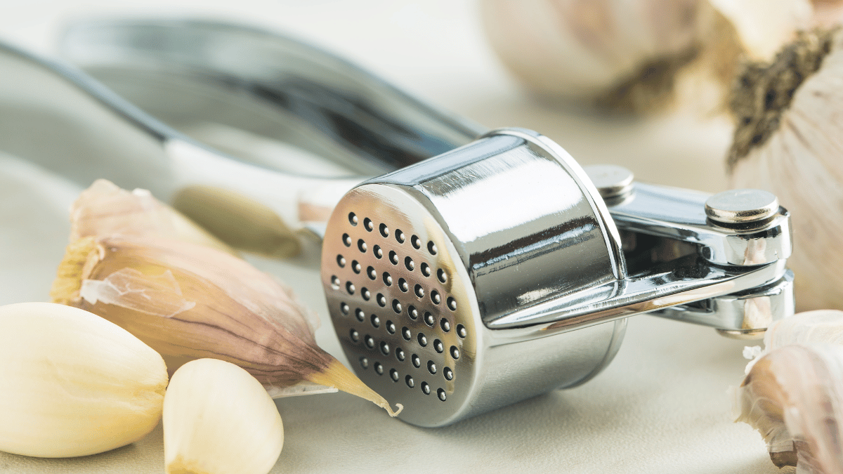 Close-up of a stainless steel garlic press surrounded by fresh garlic cloves on a light-colored surface