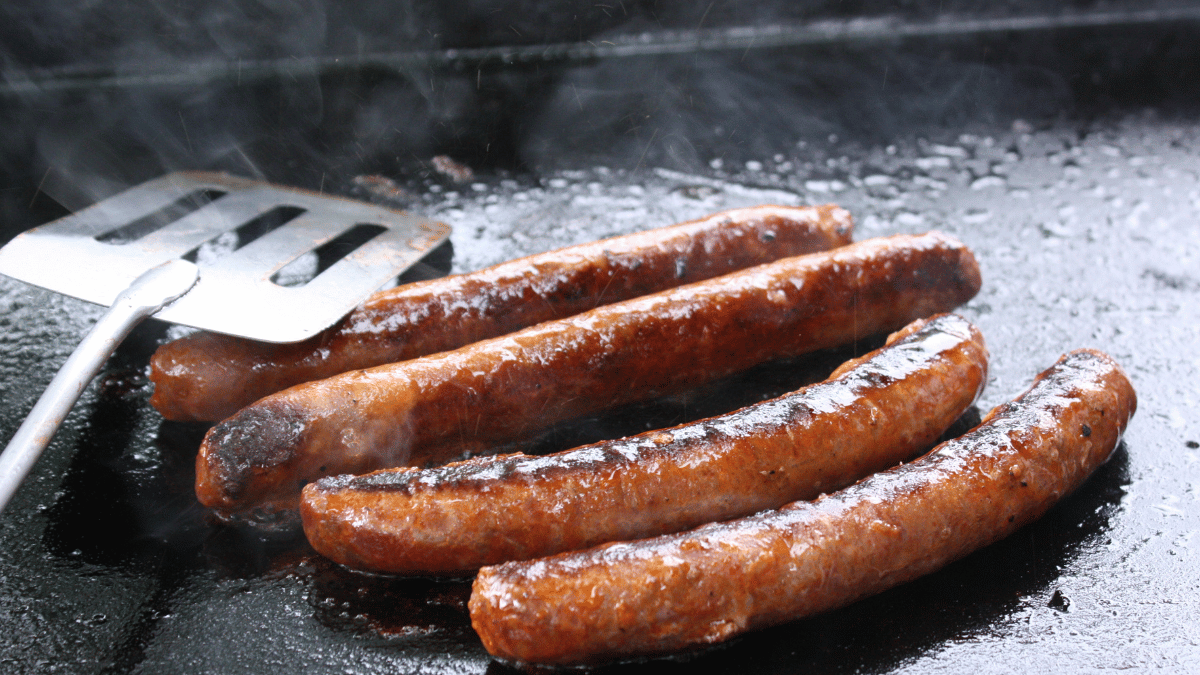 Grilled sausages cooking on a hot griddle with a metal spatula resting nearby, steam rising from the surface.