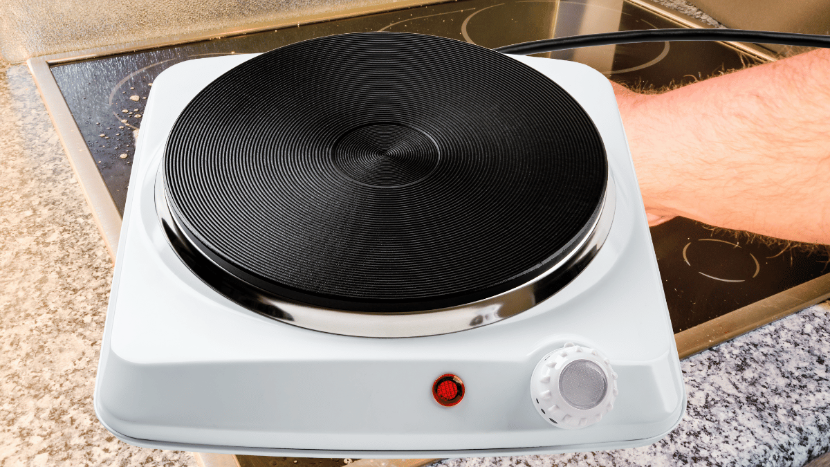 Portable electric hot plate with a single burner placed on a kitchen counter, partially covering a glass-ceramic stovetop being cleaned by hand.
