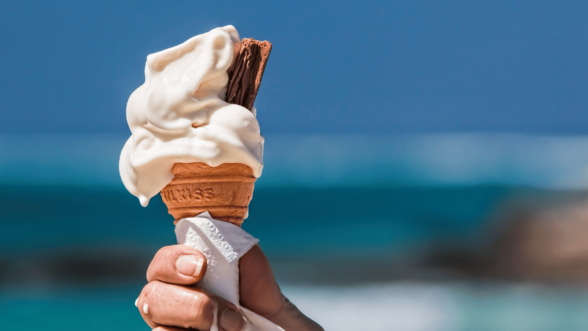 Close-up of a hand holding a melting vanilla ice cream cone with a chocolate flake against a blurred ocean background on a sunny day.