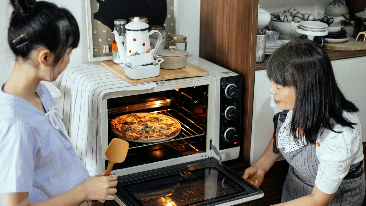 Two women in a kitchen checking a cooked pizza in a countertop oven, with one holding a spatula and the other opening the oven door.