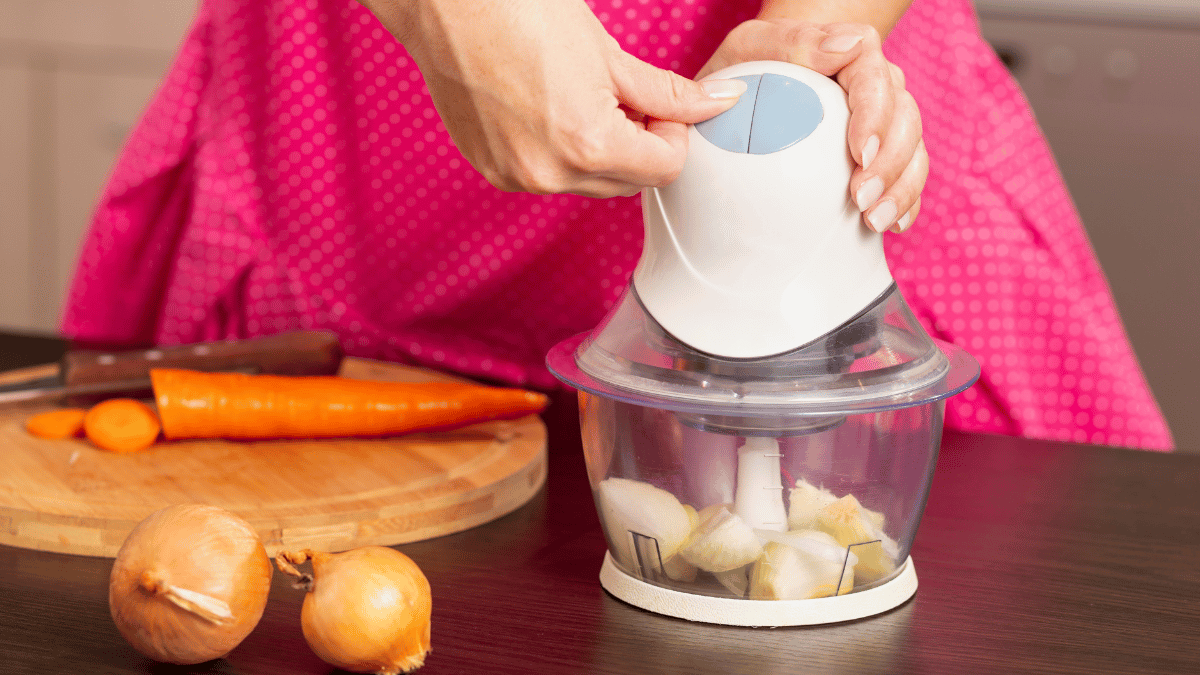 Close-up of a person using a food chopper to dice onions, with whole onions, a carrot, and a cutting board in the background on a kitchen counter.