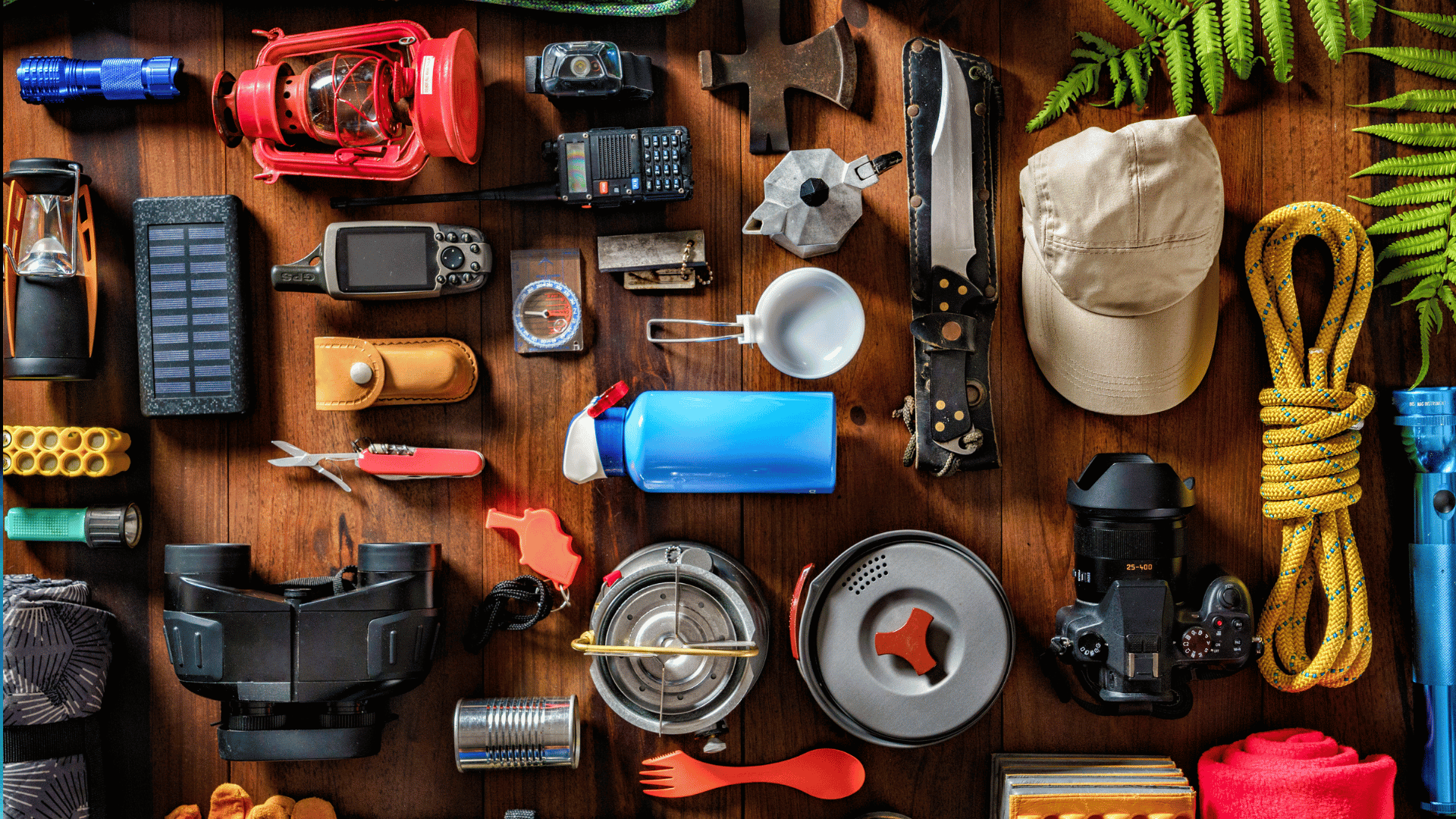 Flat lay of various camping and survival gear on a wooden surface, including lanterns, a GPS device, compass, knives, hat, rope, flashlight, water bottle, camera, cookware, and other essential tools arranged neatly.