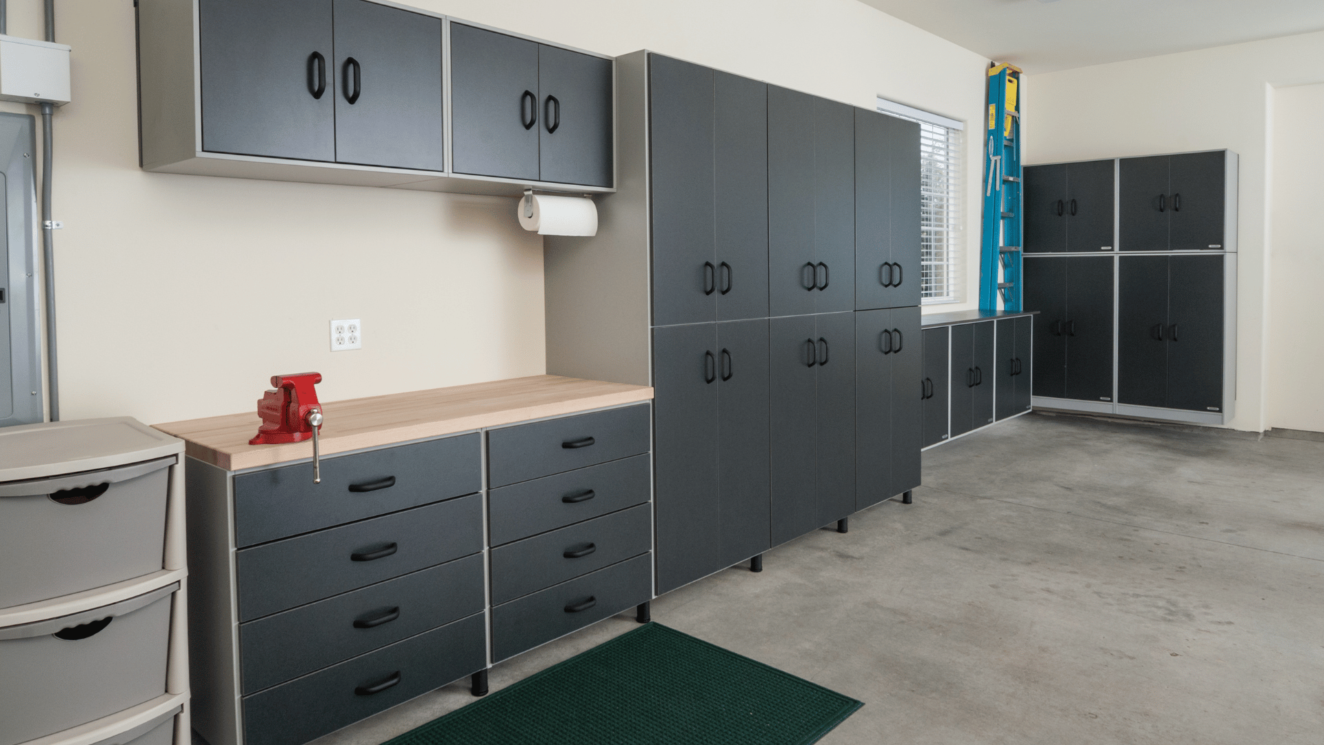 Clean and organized garage with dark gray storage cabinets, a wooden workbench, mounted overhead cupboards, a red bench vise, and a blue ladder stored in the corner.