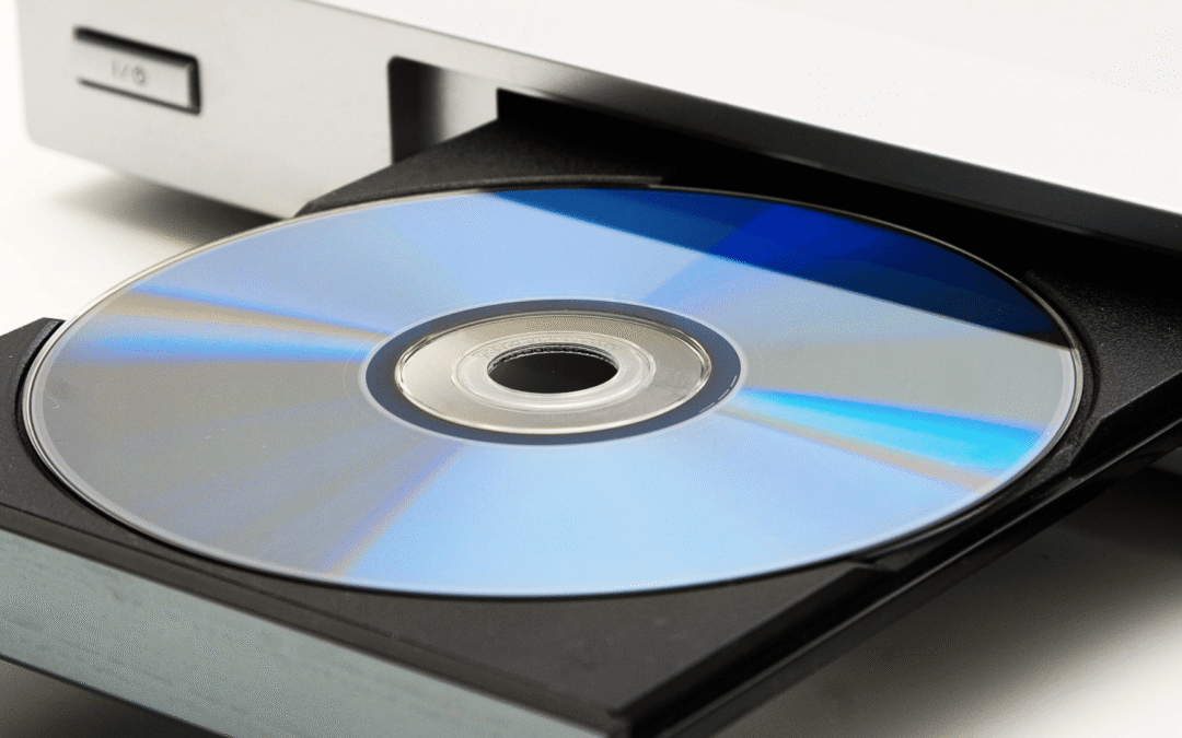 A close-up of a DVD or Blu-ray disc sitting in the tray of a silver disc drive, partially ejected, with the reflective surface of the disc showing blue and rainbow-like hues.