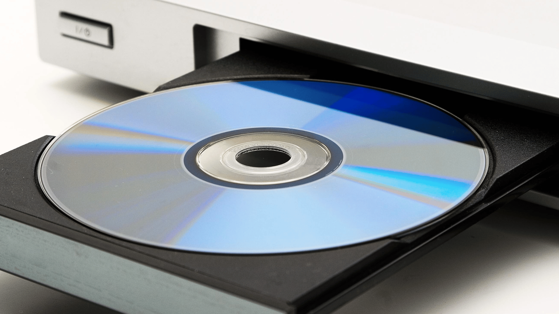 A close-up of a DVD or Blu-ray disc sitting in the tray of a silver disc drive, partially ejected, with the reflective surface of the disc showing blue and rainbow-like hues.