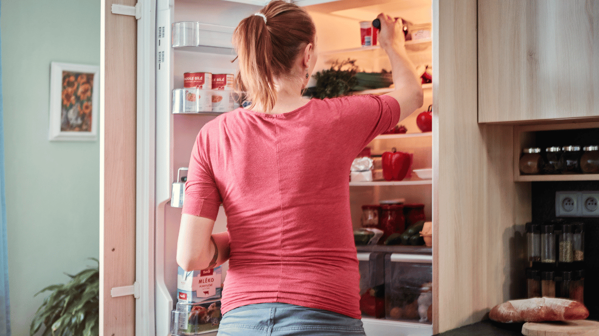 Woman in a red shirt seen from behind, reaching into a well-organized refrigerator filled with fresh produce, jars, and dairy items in a cozy kitchen setting.