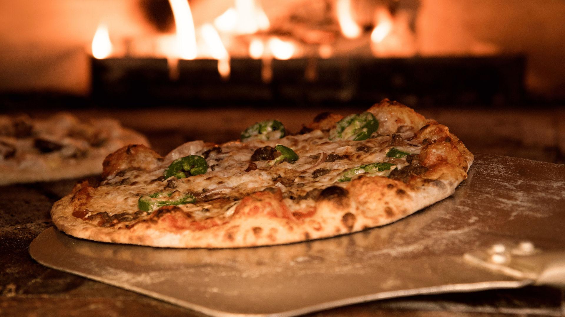 Wood-fired pizza topped with cheese, sausage, and sliced green peppers being pulled from a traditional pizza oven with flames glowing in the background.