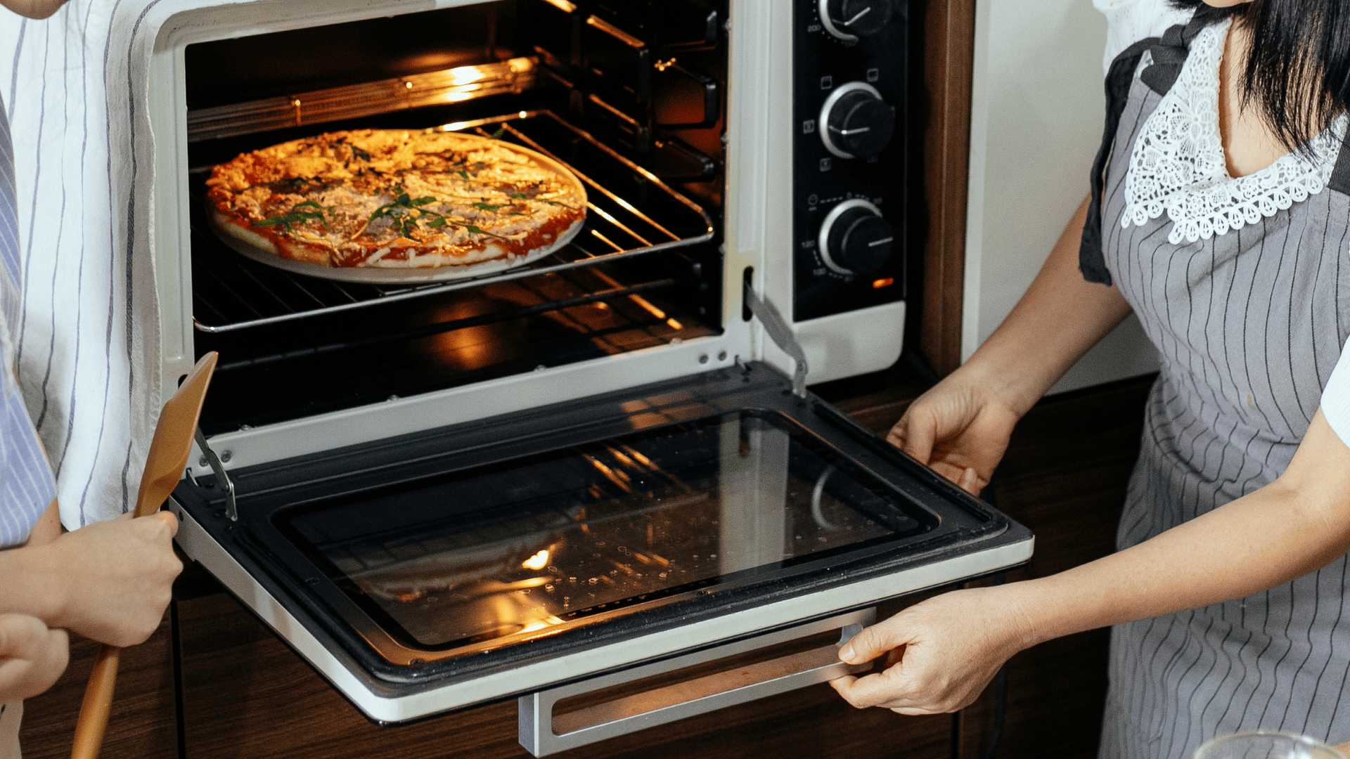 Two women baking a homemade pizza in a countertop oven, with one opening the oven door and the other holding a wooden spatula.