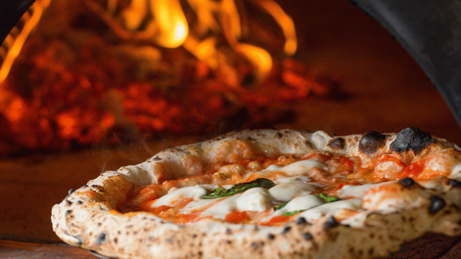 Neapolitan-style pizza baking in a wood-fired brick oven with visible flames and glowing embers in the background.