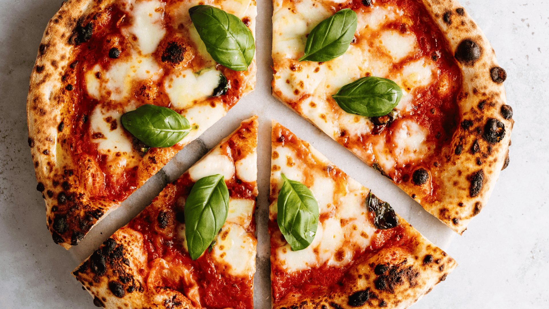 Overhead view of a sliced Margherita pizza with melted mozzarella, fresh basil leaves, and charred crust on a light stone surface.