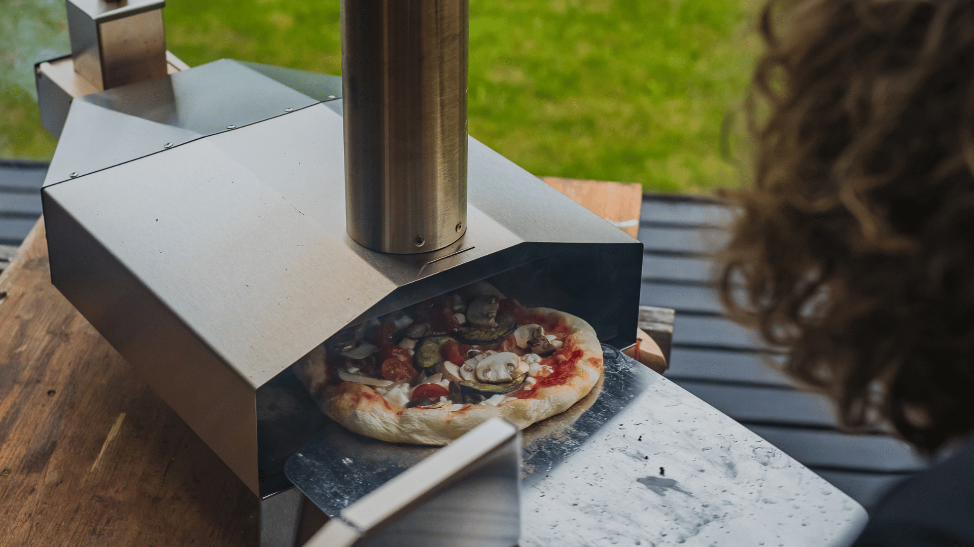 A freshly topped homemade pizza with mushrooms and tomatoes being placed into a stainless steel outdoor pizza oven on a wooden table, with a person observing from the side and a grassy yard in the background.