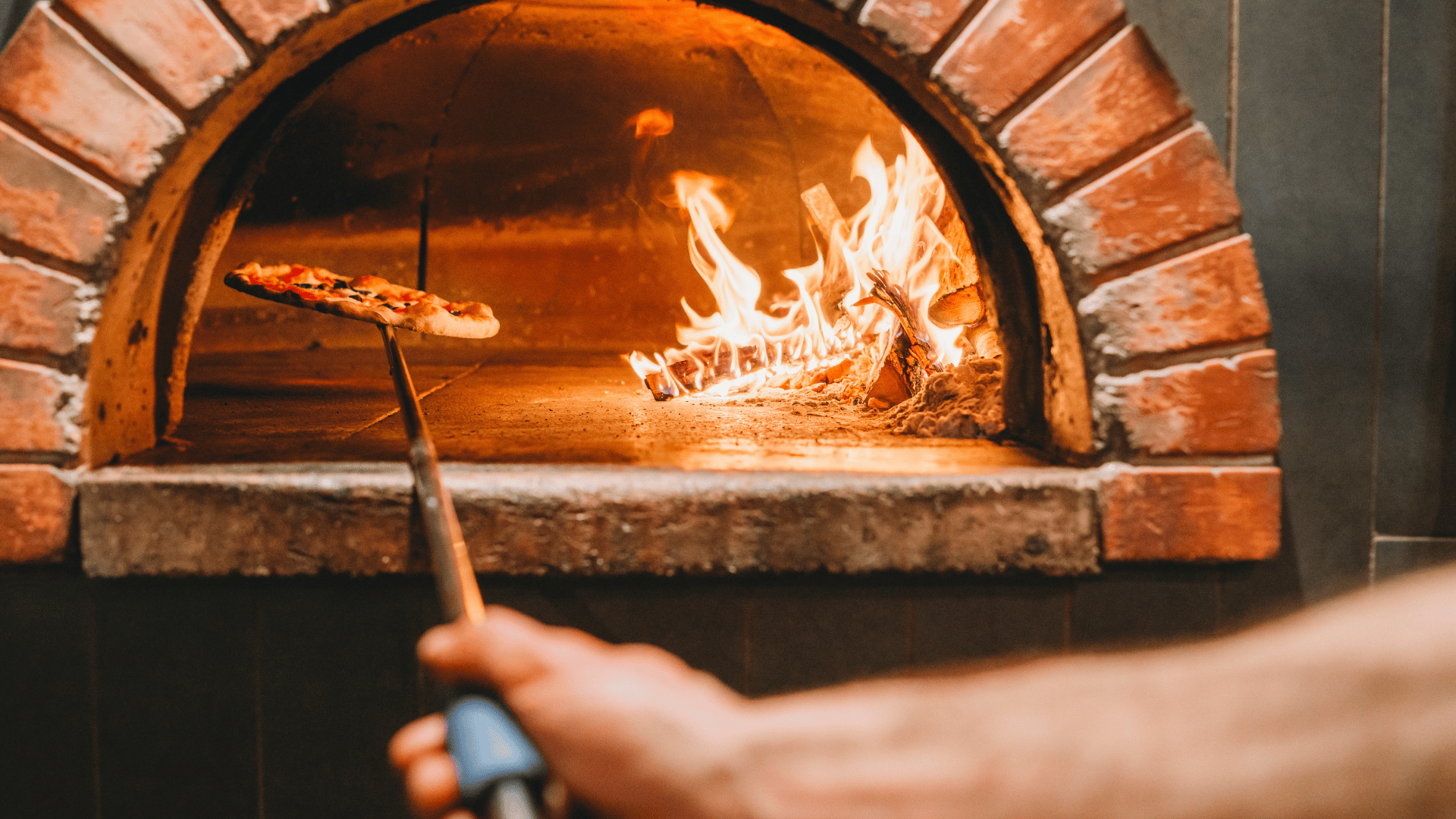 A hand uses a pizza peel to place a pizza inside a traditional brick wood-fired oven with a blazing fire burning on one side, creating an authentic pizzeria atmosphere.