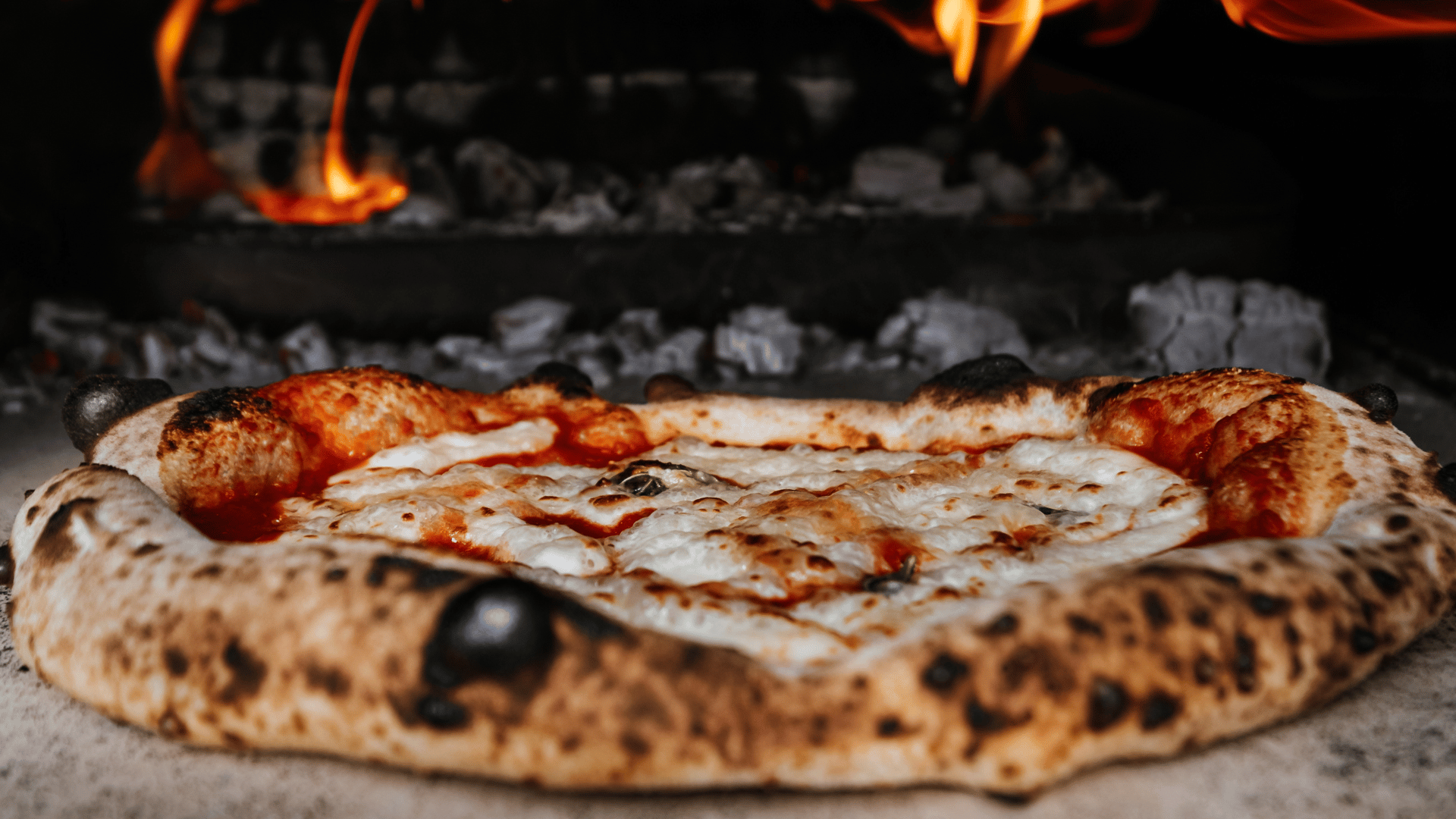 Close-up of a Neapolitan-style pizza with charred crust and melted mozzarella cheese baking directly on the stone surface of a wood-fired oven, with glowing embers and flames in the background.