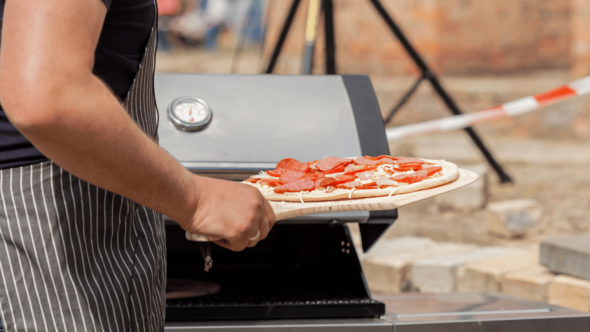 Person in a striped apron placing a raw pepperoni pizza into an outdoor grill-style pizza oven using a wooden pizza peel, with a thermometer visible on the oven lid.