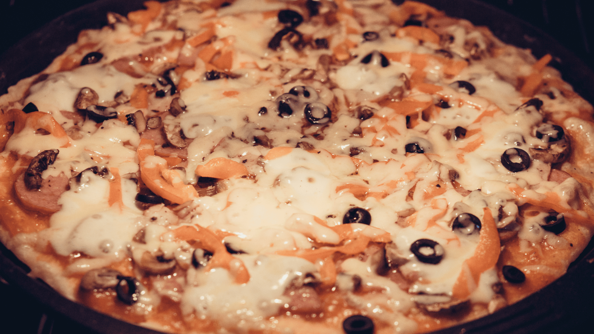 Close-up of a freshly baked pizza topped with melted cheese, sliced black olives, orange bell peppers, mushrooms, and sausage on a dark baking tray.