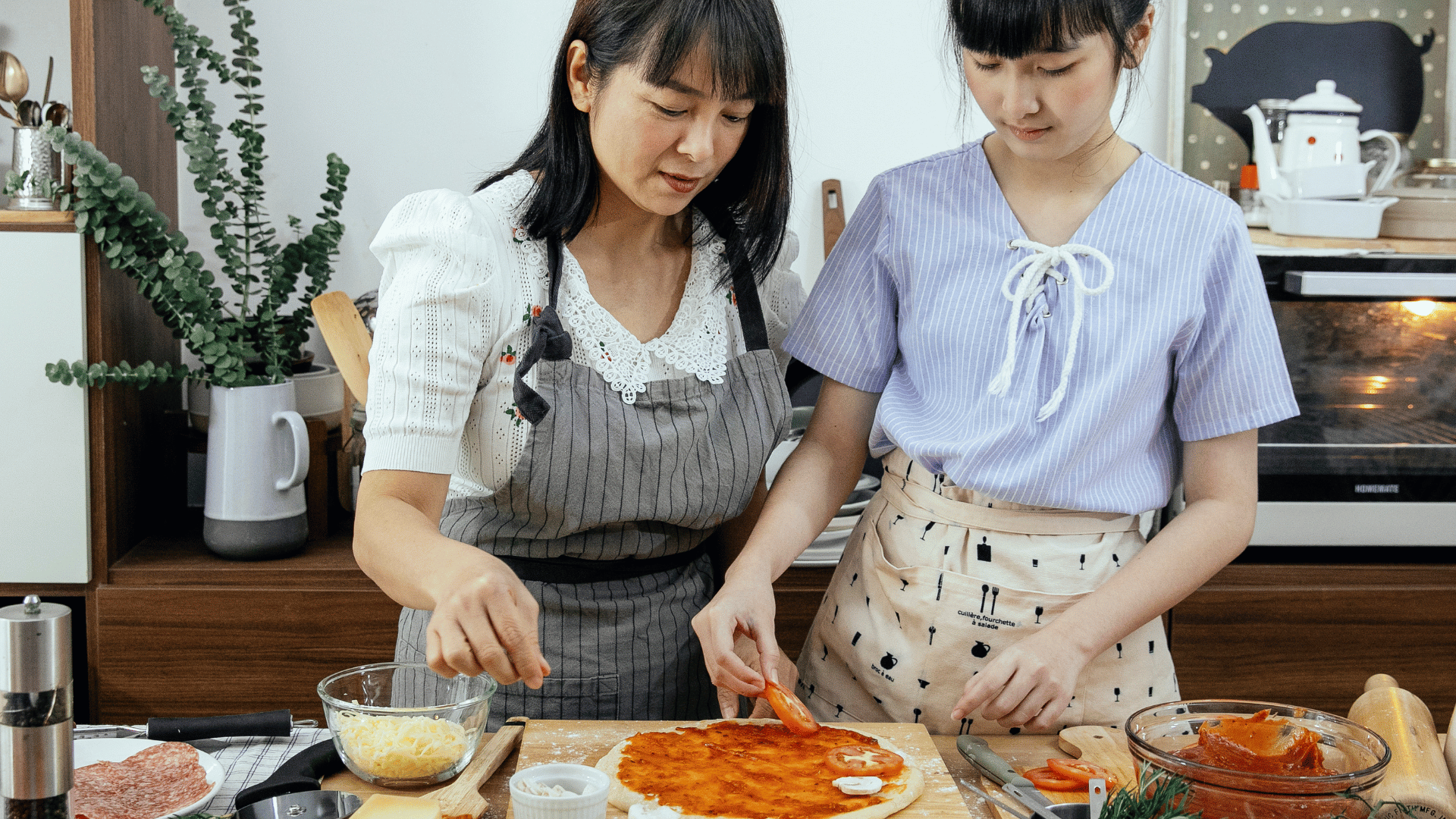 Two women in aprons preparing a homemade pizza in a kitchen, carefully placing sliced tomatoes on a pizza base already covered with tomato sauce. Various ingredients and kitchen tools are arranged around them on the countertop.