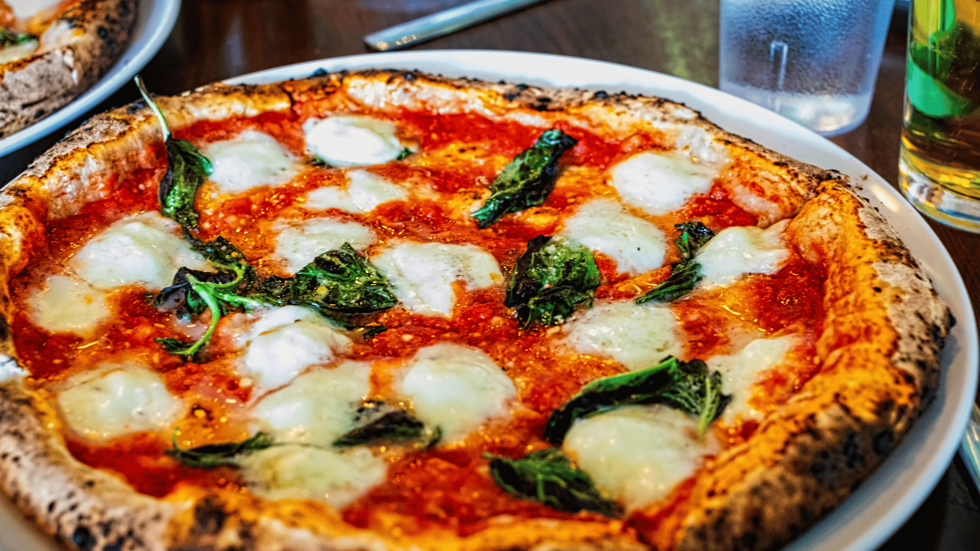 A freshly baked Neapolitan-style Margherita pizza served on a white plate, topped with melted mozzarella, vibrant basil leaves, and a rich tomato sauce, with a lightly charred crust.