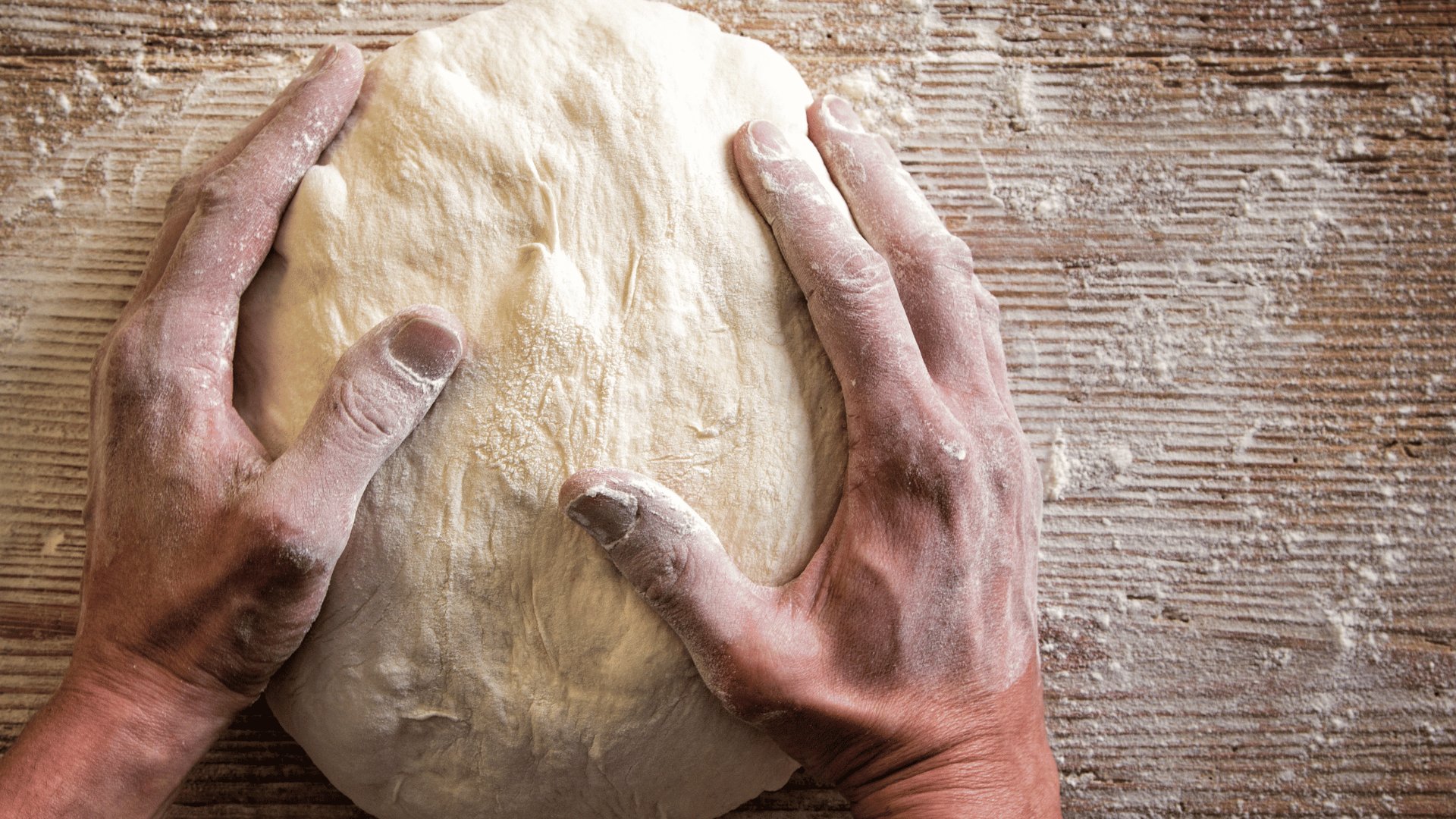 Close-up of flour-covered hands shaping a ball of pizza dough on a rustic wooden surface lightly dusted with flour.