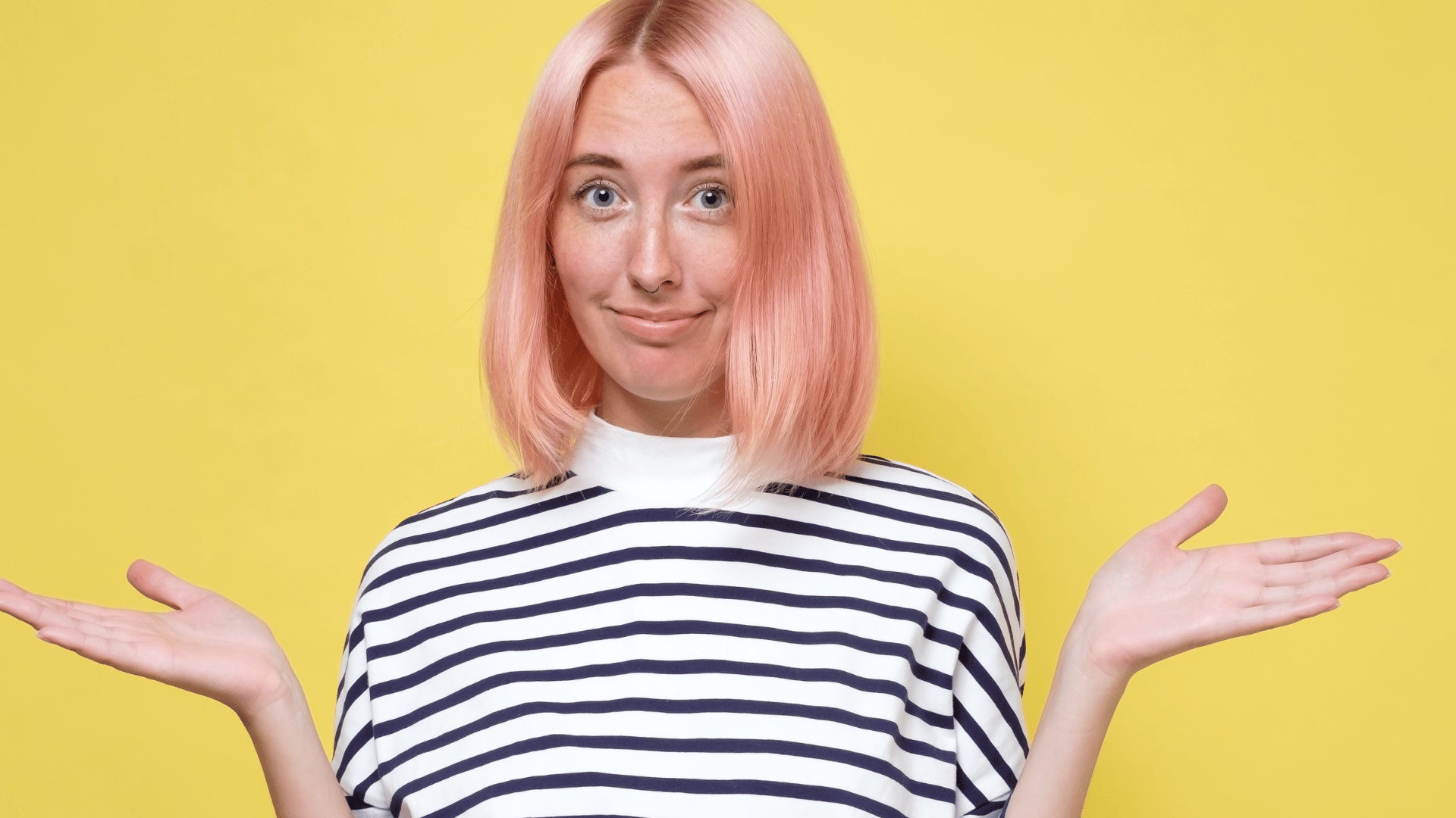 Person with pink hair wearing a striped shirt shrugging with both hands raised, standing in front of a bright yellow background and expressing uncertainty or indifference.