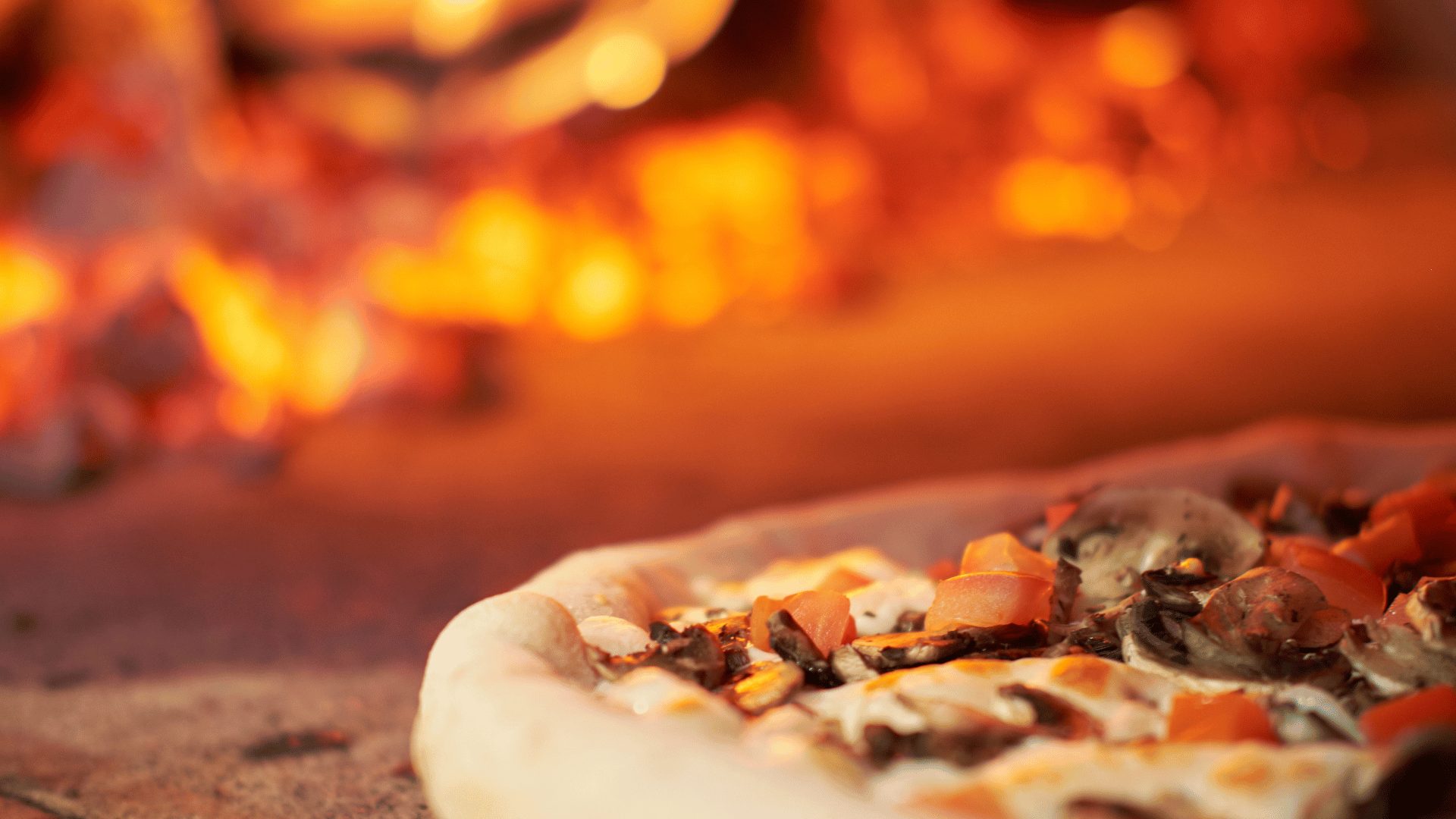 Close-up of a pizza with mushrooms, cheese, and diced tomatoes baking in a wood-fired oven, with glowing embers and flames in the blurred background.