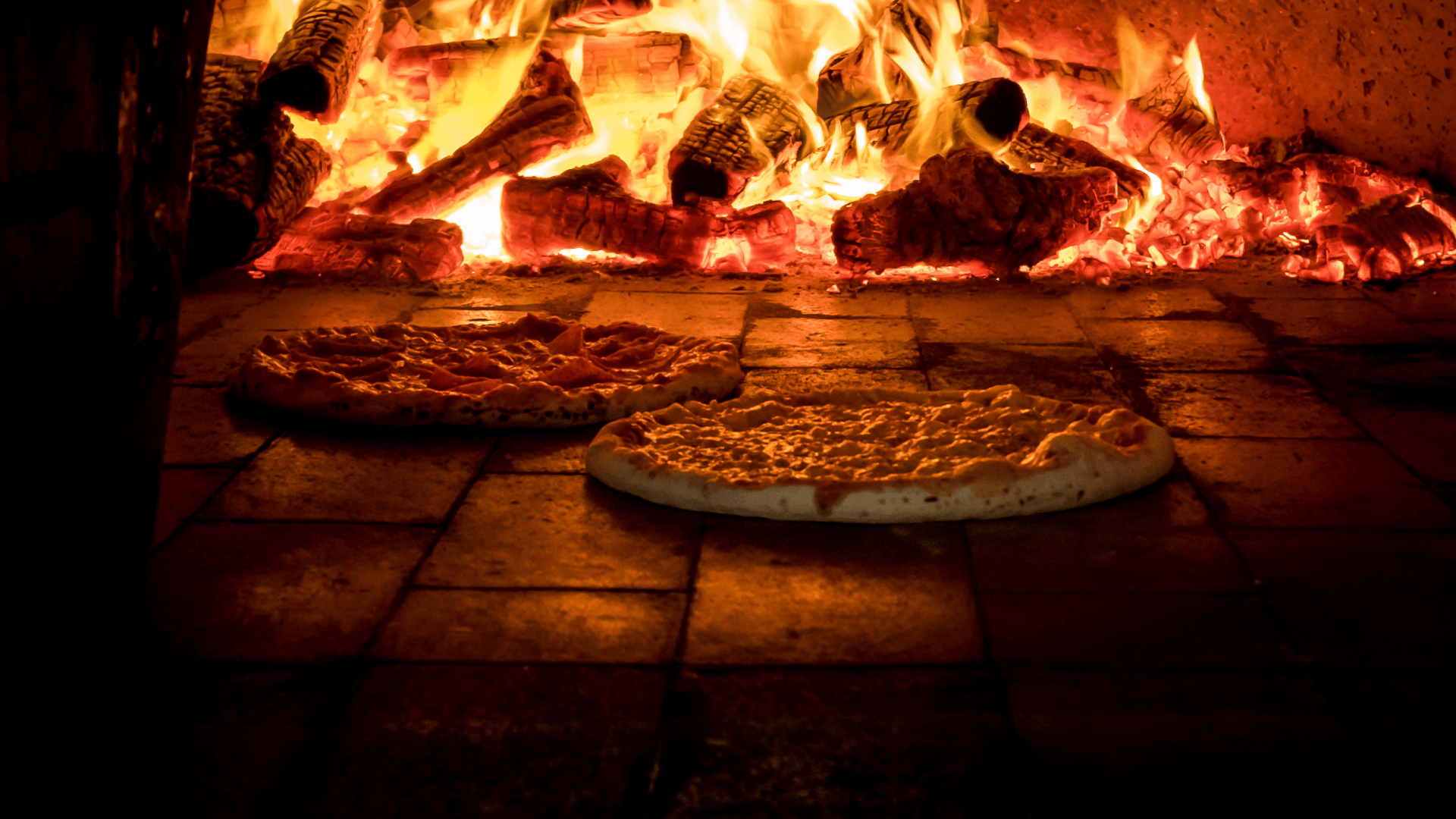 Two pizzas baking directly on the stone floor of a traditional wood-fired oven, with large flames and glowing embers in the background.