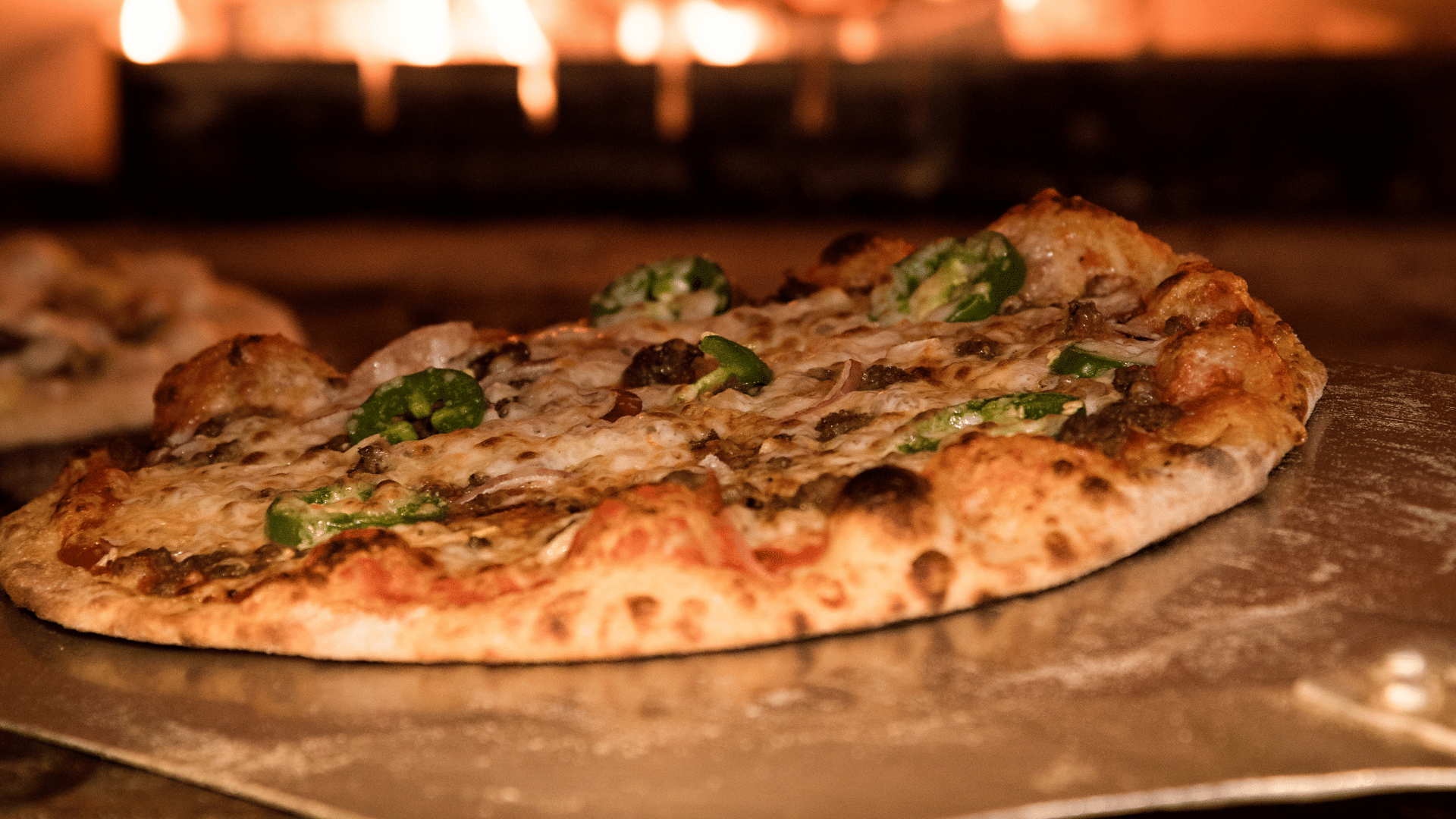 Close-up of a freshly baked pizza topped with cheese, sausage, and sliced jalapeños being pulled from a wood-fired oven on a metal pizza peel, with flames glowing in the background.