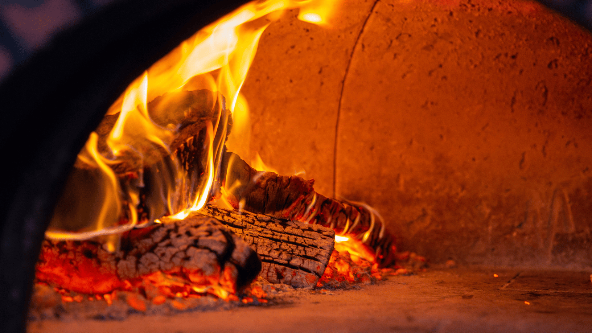Close-up view of a roaring fire with burning wood inside a traditional dome-shaped brick pizza oven, creating intense heat for baking.