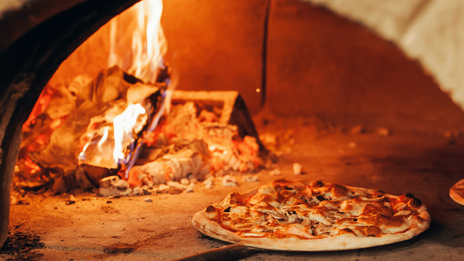 A wood-fired pizza oven with a flame burning on one side and a pizza baking directly on the oven floor, surrounded by glowing embers and rustic stone interior.