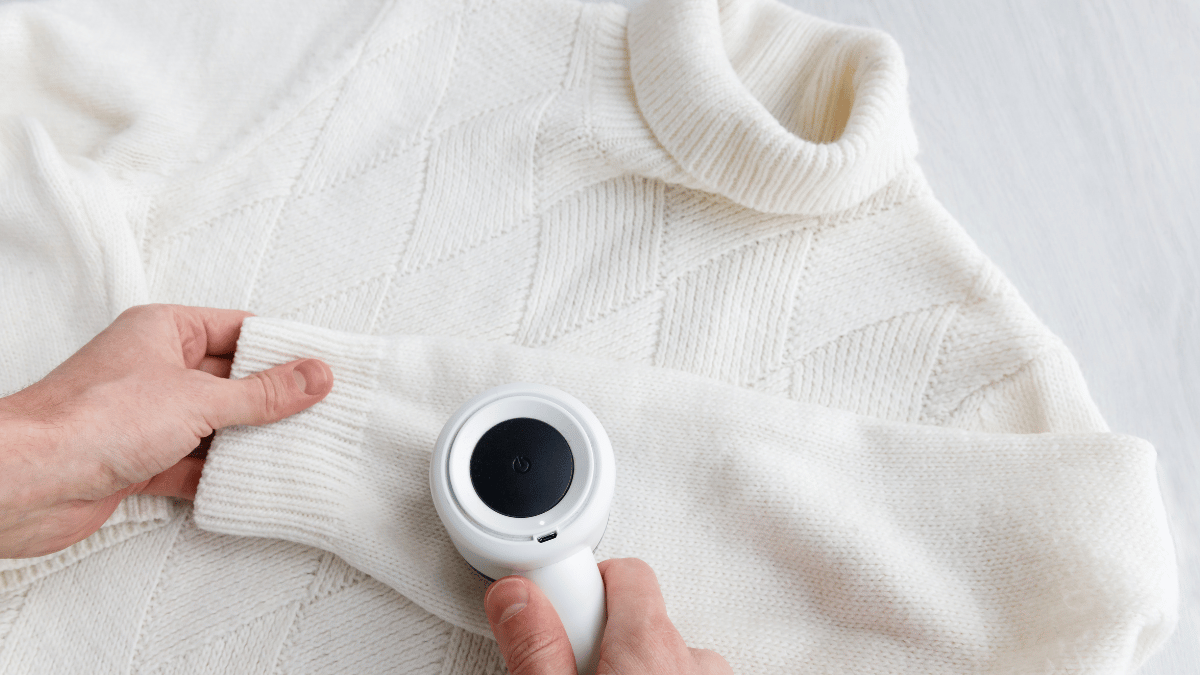 Person using a fabric shaver or lint remover on the sleeve of a cream-colored knitted sweater laid flat on a light surface.
