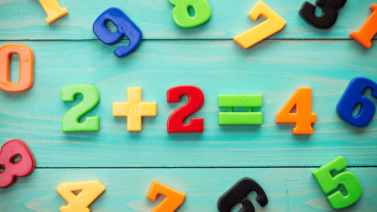 Colourful plastic numbers and math symbols on a turquoise wooden surface form the equation "2 + 2 = 4", surrounded by scattered digits in various colours.
