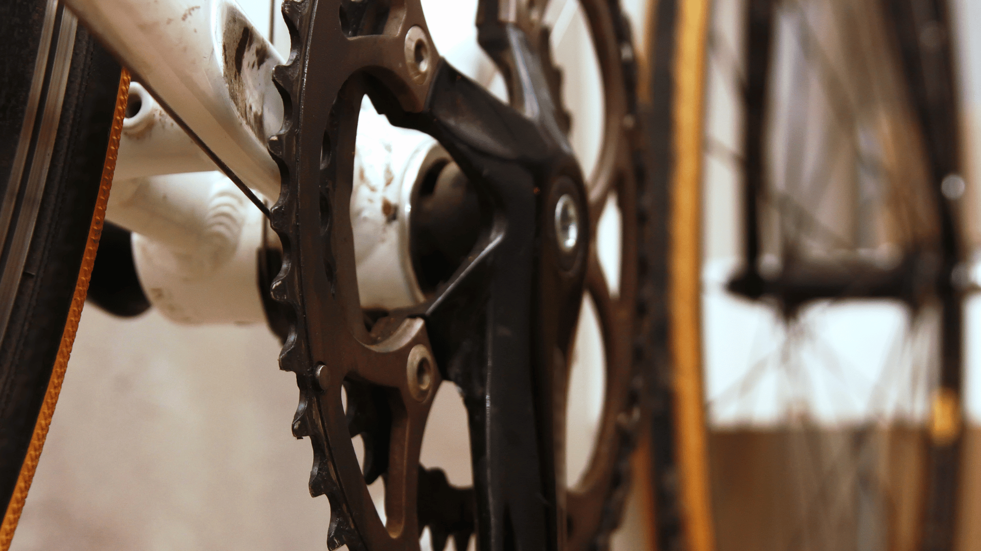 Macro view of a bicycle’s drivetrain showing a weathered metal chainring and black crank arm attached to a white frame, with a blurred yellow-striped tire and spokes in the background.