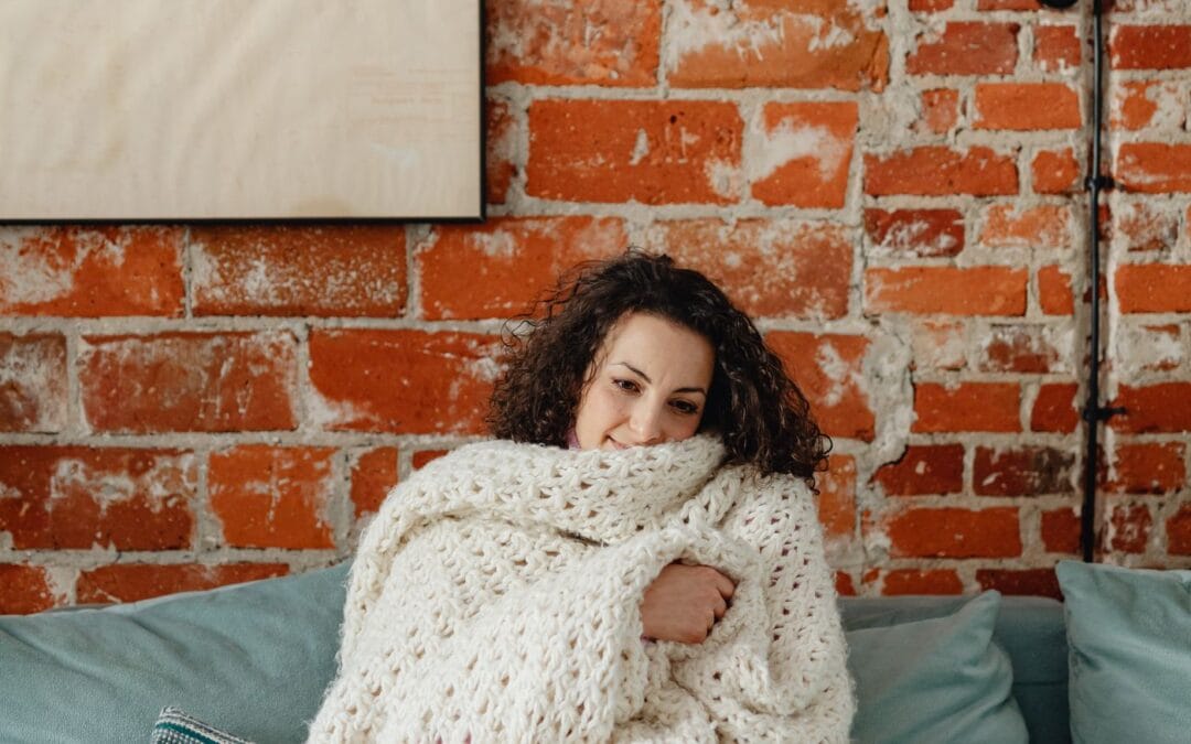 Person sitting on a sofa wrapped in a thick cream knitted blanket, illustrating heated throws and electric blankets