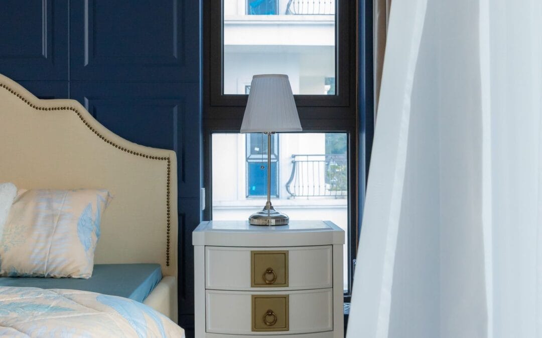 A lamp on a bedside table next to a made bed, with daylight coming through the bedroom window
