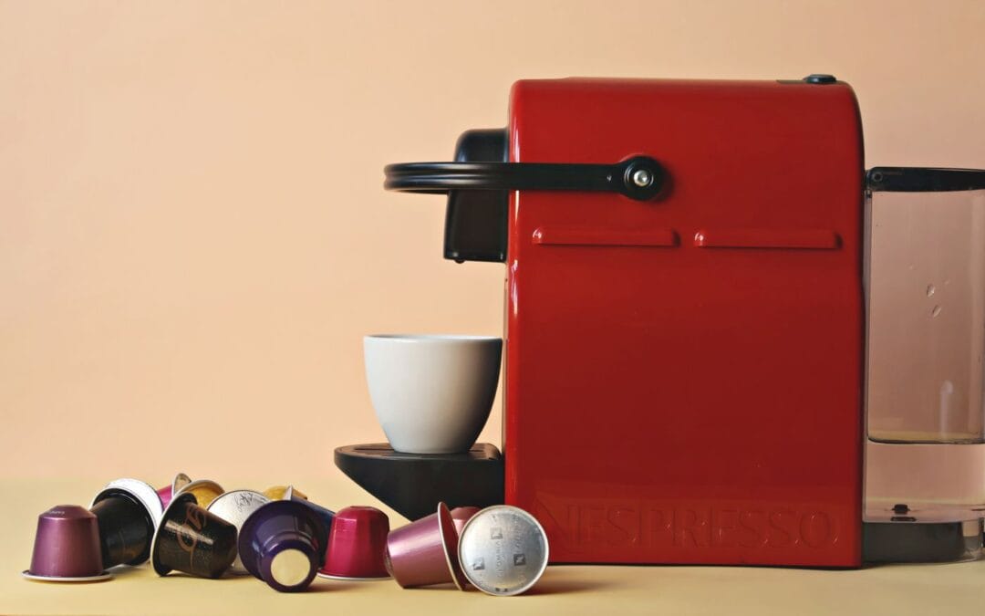 Red compact espresso machine with a white mug under the spout and coffee capsules scattered on a pale counter.