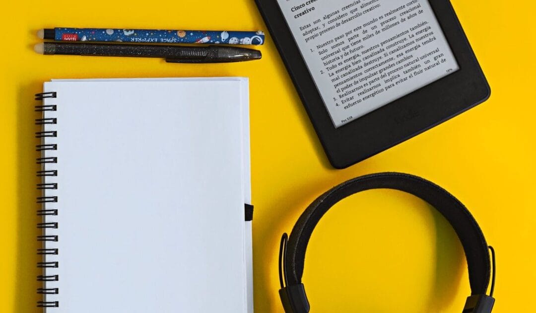 An e-reader on a yellow background beside a notebook and headphones