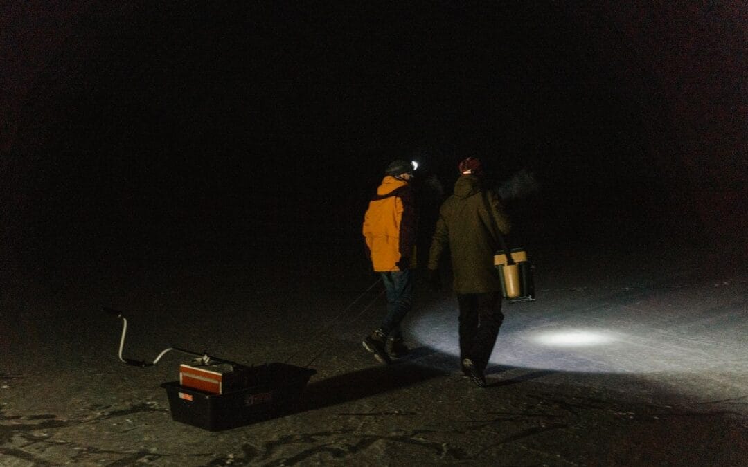 Two people walking in the dark, one wearing a lit head torch casting a beam on the ground ahead