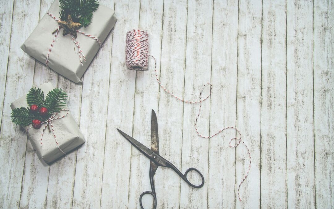 Holiday gift-wrapping scene: silver presents tied with red-and-white twine, pine sprigs and berries on a white wooden table, plus scissors and a string spool nearby.