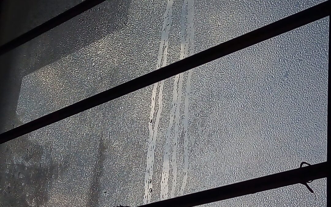 Frosted glass window with vertical water streaks and diagonal black metal bars crossing the frame.
