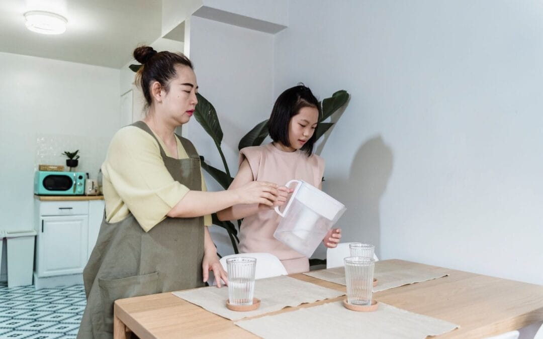 A woman and a child pouring filtered water from a white filter jug into glasses on a kitchen table